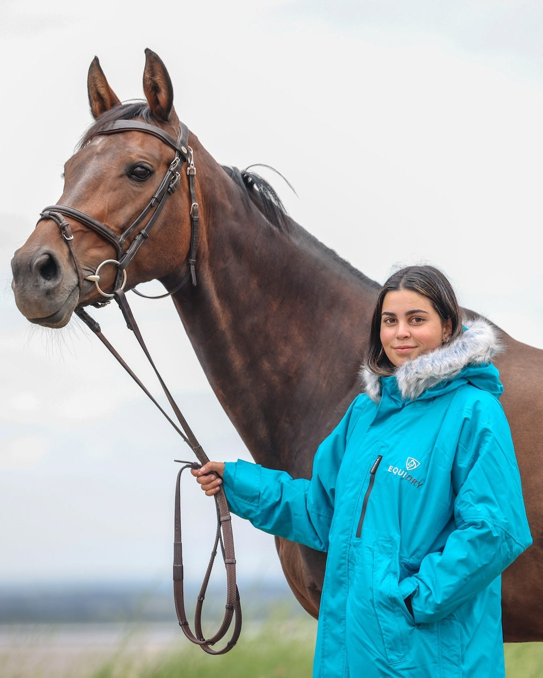 EQUIDRY Evolution Lux Women's Warm Waterproof Winter Horse Riding Coat Thick Fleece Lining with Faux Fur Trim Hood in turquoise rider stood with horse