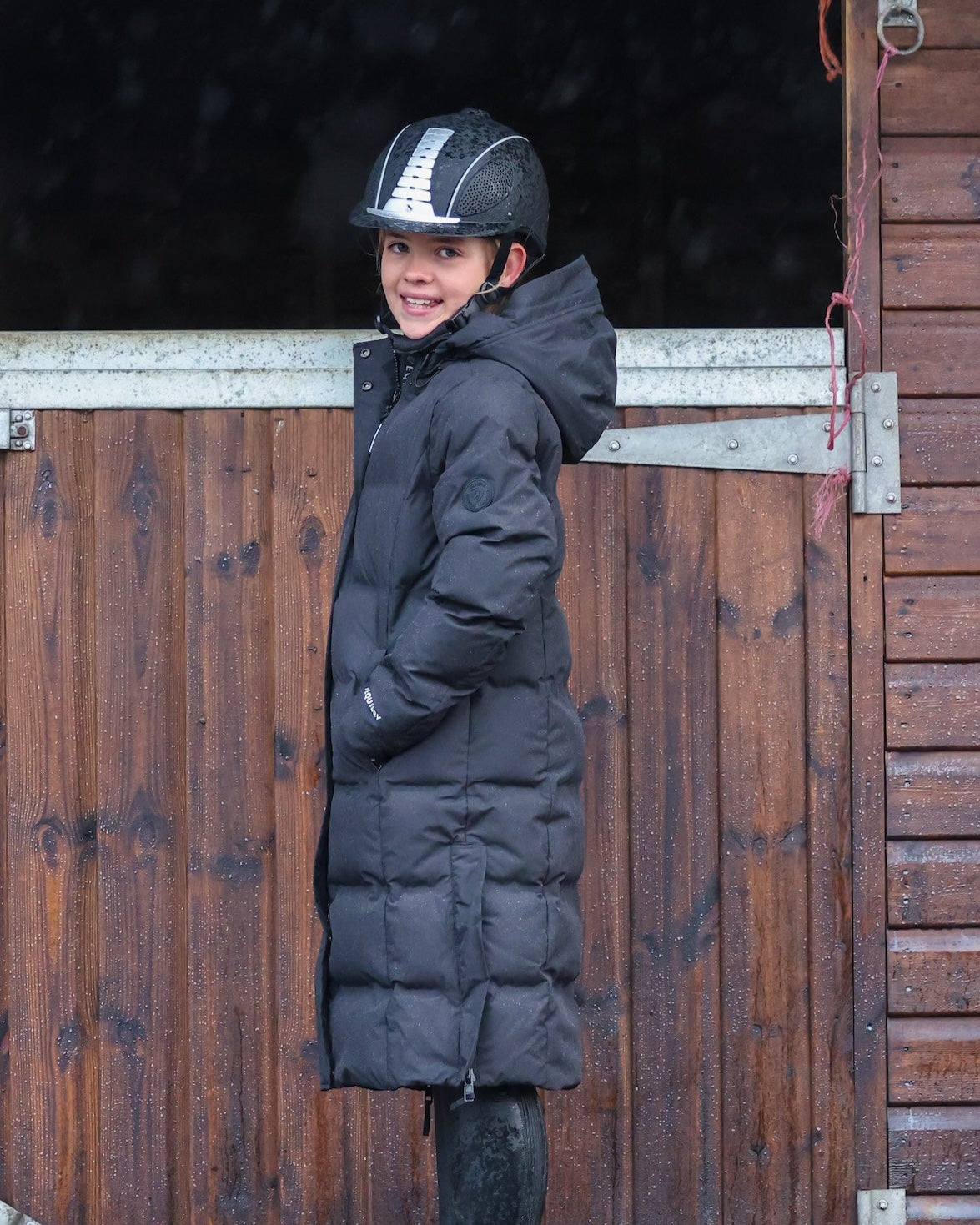 Girl in EQUIDRY Thora Black Waterproof Riding Puffer Coat & Equestrian Helmet at Stable