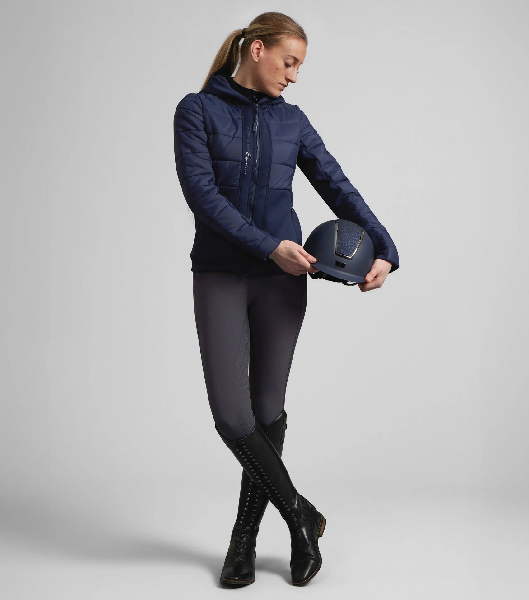 Premier Equine Agata Ladies Riding Jacket Navy: Equestrian woman holding helmet. Quilted, hooded riding jacket for women.