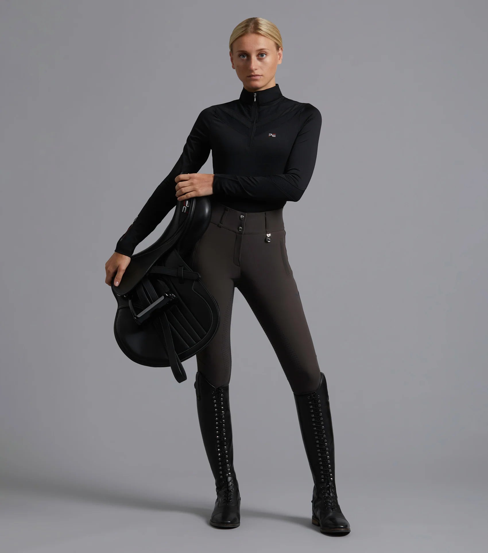 Premier Equine Arclos Black Riding Top. Equestrian Apparel, ladies technical long sleeve. Equestrian model blonde woman holding saddle.