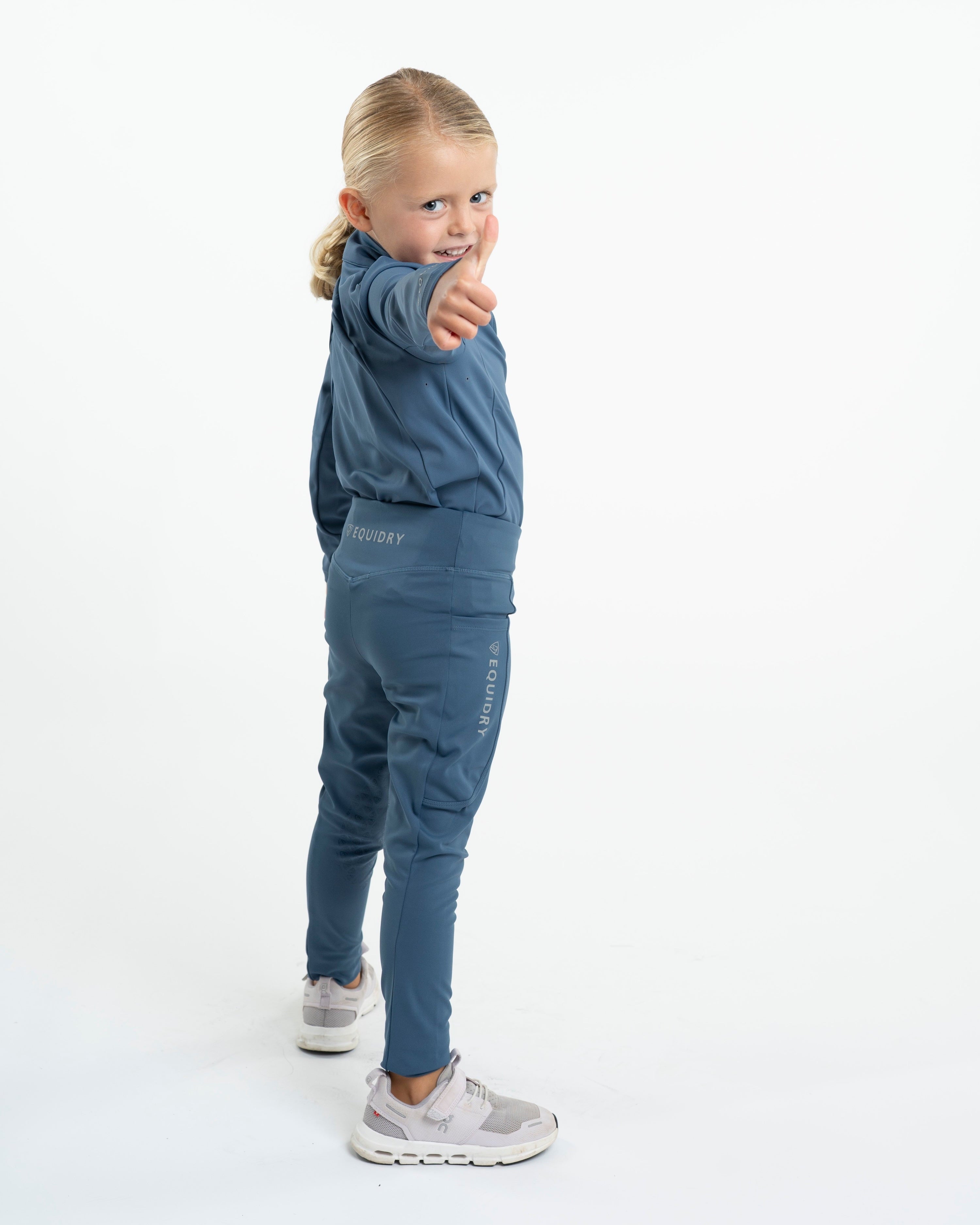 Equidry: Young girl in steel blue riding leggings giving a thumbs-up. Water-repellent knee grip leggings for kids. -…