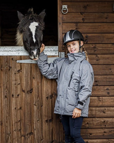 Shortie Child's Oversized Short Waterproof blouson style Riding Jacket Thin Fleece Lining Stowaway Hood in charcoal/penelope pink girl stood with horse