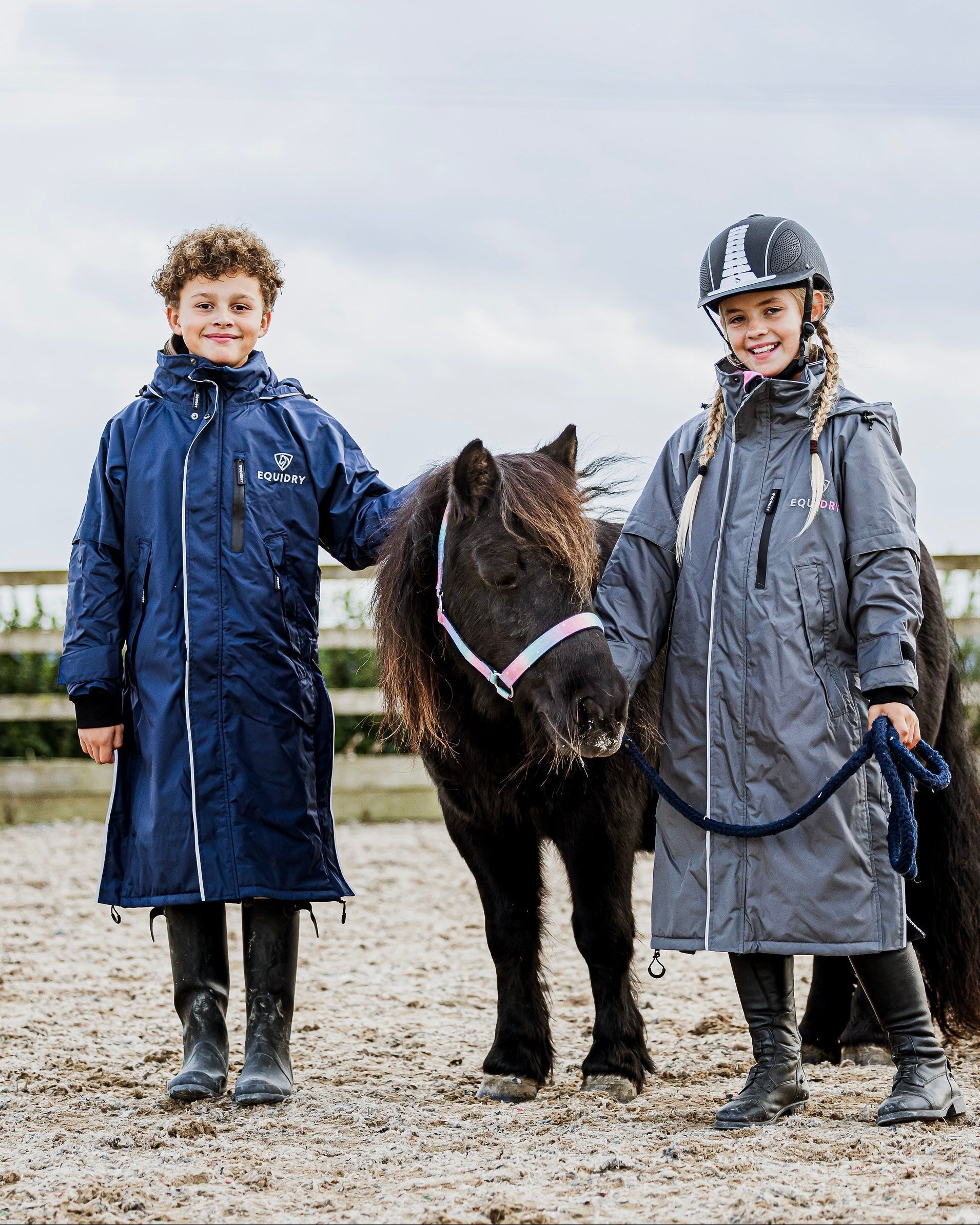 EQUIDRY waterproof horse riding raincoat thin fleece pro ride reflective in navy and charcoal/pale pink children's kids stood in arena with shetland