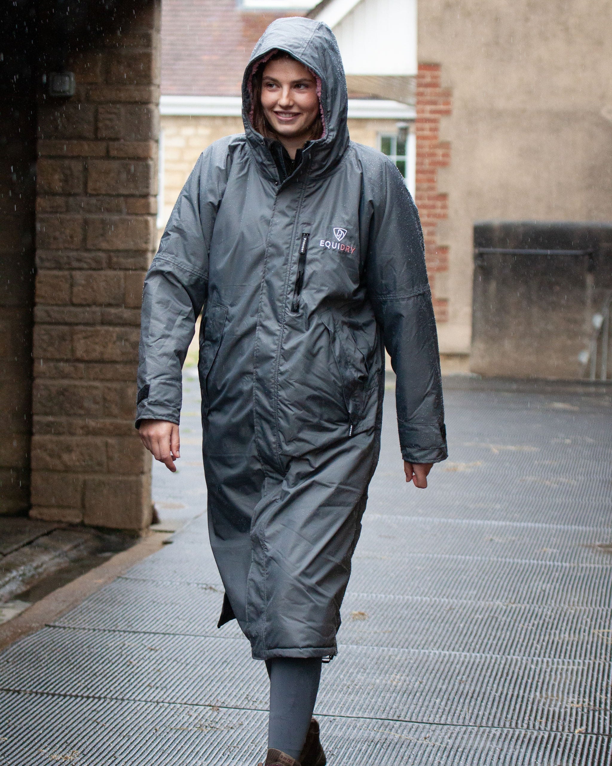 EQUIDRY women's long waterproof horse riding coat in charcoal/pale pink showing groom on yard sheltering from the rain