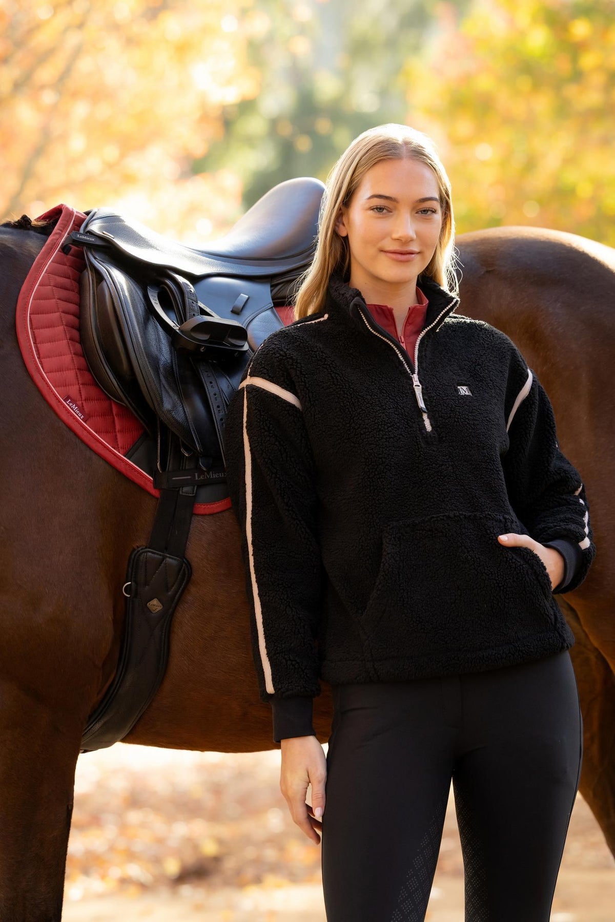 Woman in black LeMieux Avery Teddy Quarter Zip Fleece posing with a horse and saddle. Equestrian style.