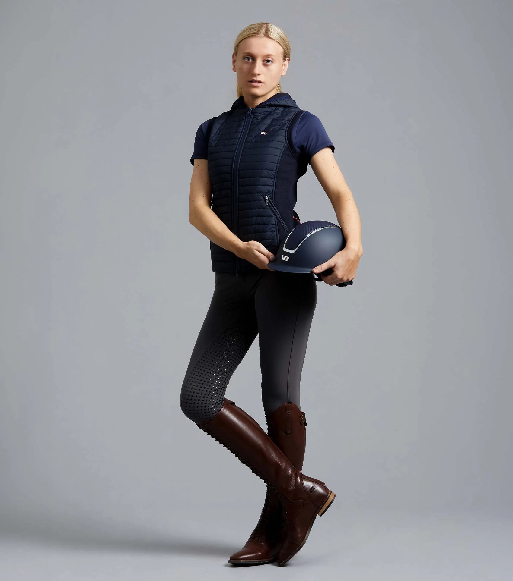 Equestrian woman wearing PREMIER EQUINE navy riding gilet and brown boots holding helmet.