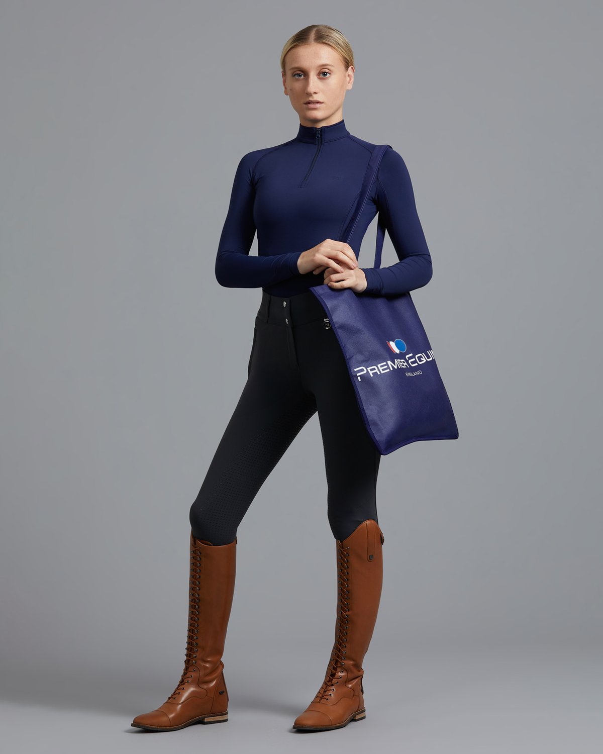 Premier Equine Navy Tote Bag Equestrian Fashion Model. Full body shot in riding boots, breeches, and top.