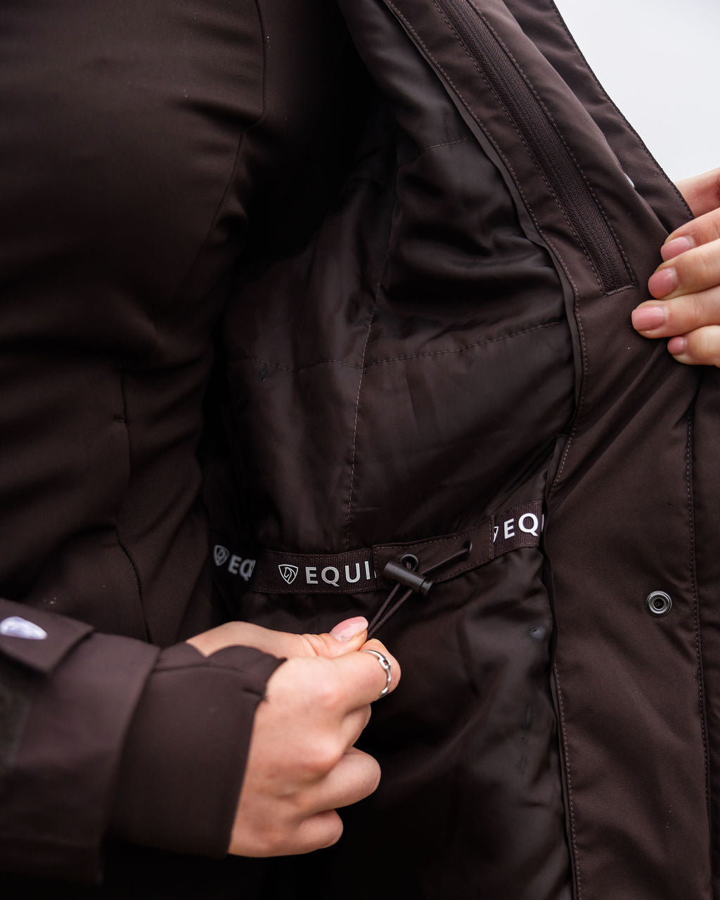 Close-up of a person adjusting the waistband of a dark brown jacket with 'EQUIDRY' branding.