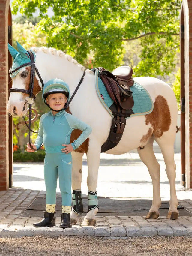 Children's equestrian apparel: Girl in LEMIEUX Lagoon riding breeches with horse, matching set, helmet, and horse accessories.