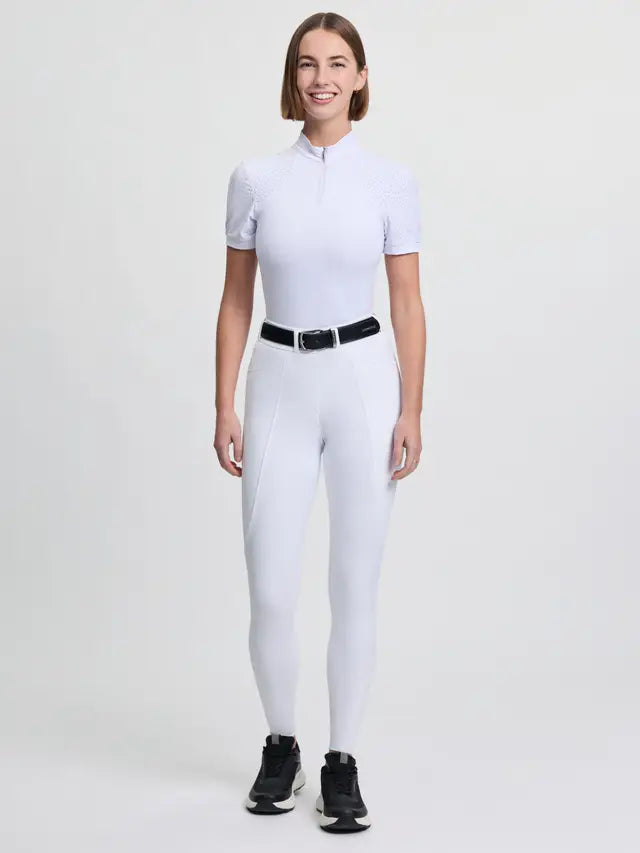 Woman wearing white LeMieux Brilliance Knee Grip Breeches and short-sleeved riding top, black belt, and black sneakers.