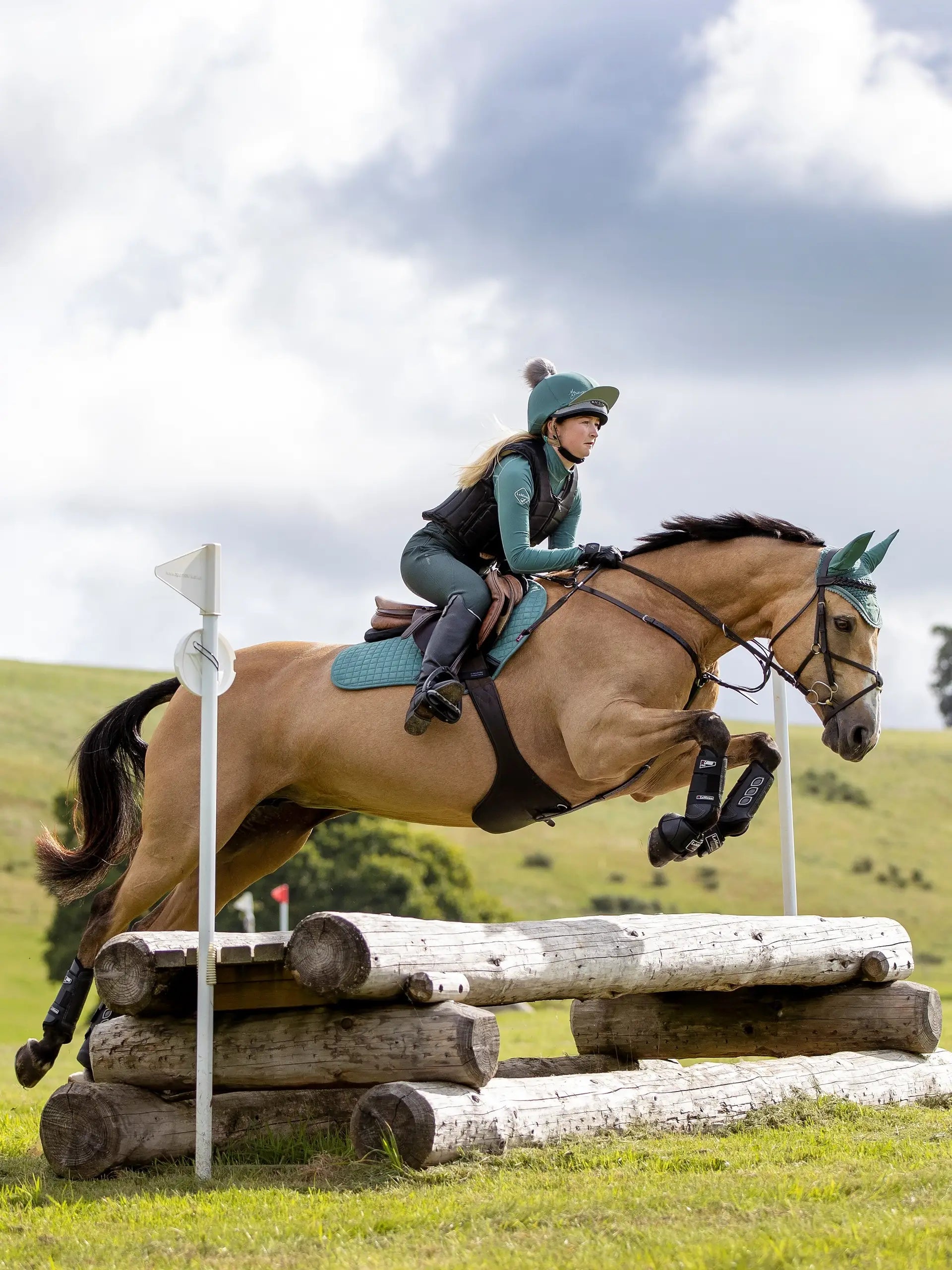 Equestrian: Horse jumping over logs. Rider in LEMIEUX Sage hat silk by Equiflair. Horse eventing action shot.