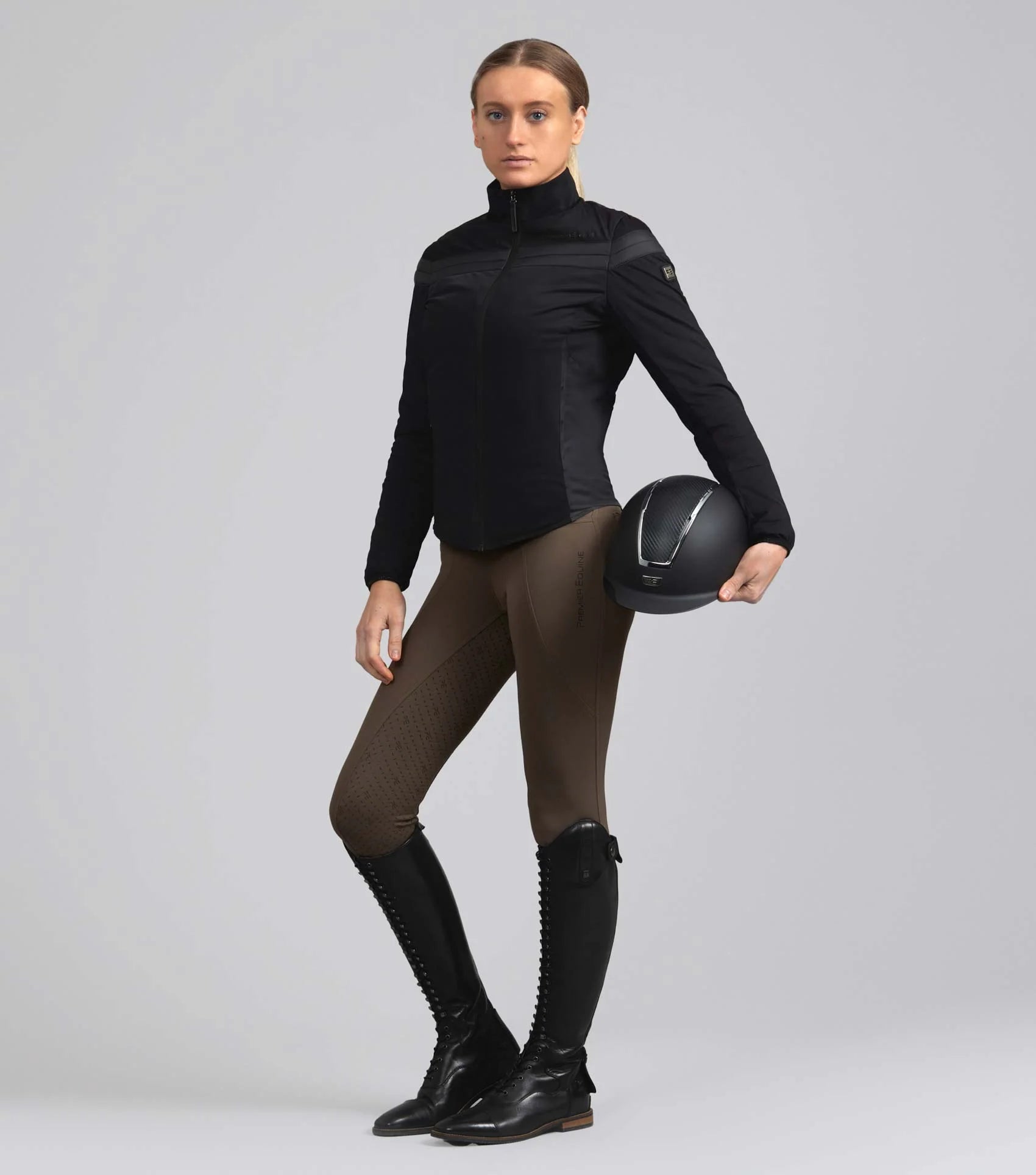 Woman in black Premier Equine Vivace riding jacket, brown breeches, boots, holding helmet. Equestrian riding gear, women's apparel.