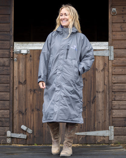 EQUIDRY Evolution Women's Fleece Coat: Stylish blonde smiles wearing grey Equidry waterproof equestrian riding coat in front of horse stable.