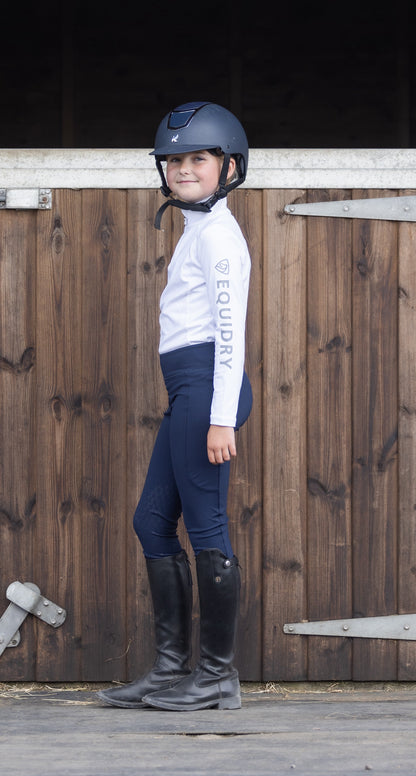 Girl in Equidry equestrian riding apparel: white base layer, navy riding leggings, boots, and helmet. Children's riding outfit.