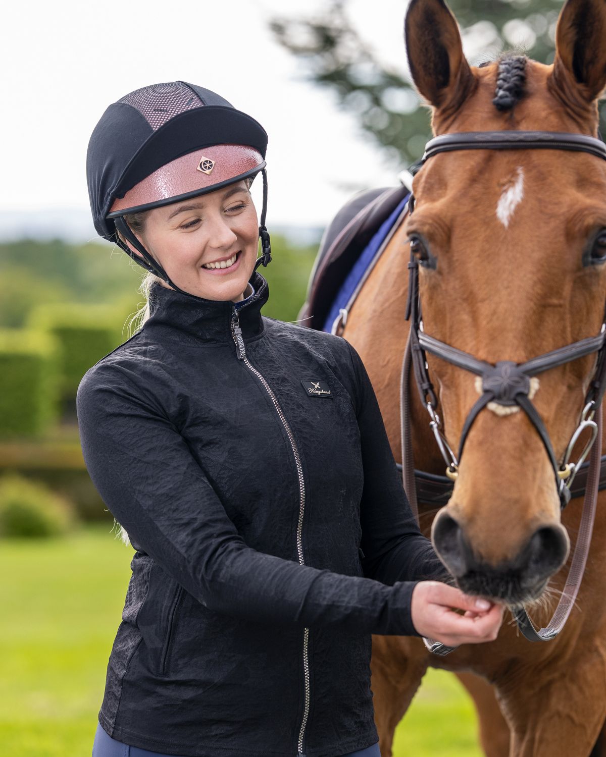 Woman feeding horse wearing Charles Owen Pro II Plus rose gold helmet and black riding jacket. Equestrian lifestyle image.
