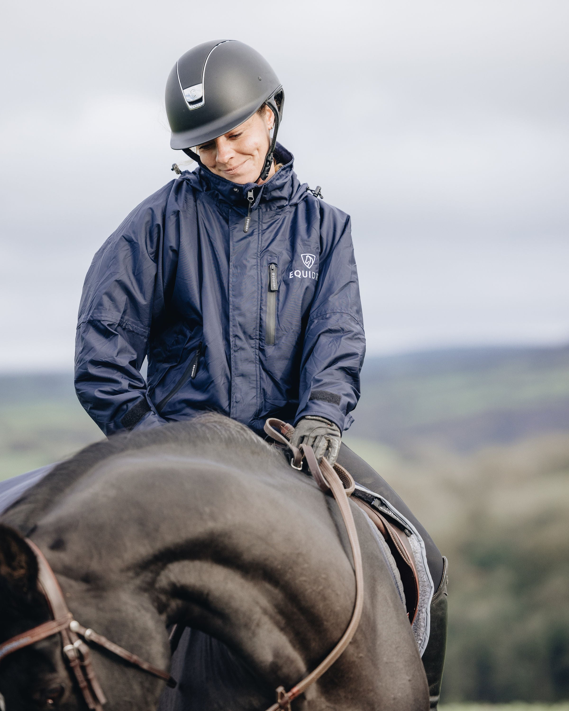 Equestrian in Navy Equidry Waterproof Riding Jacket on Horseback. Women's Shortie style with helmet.