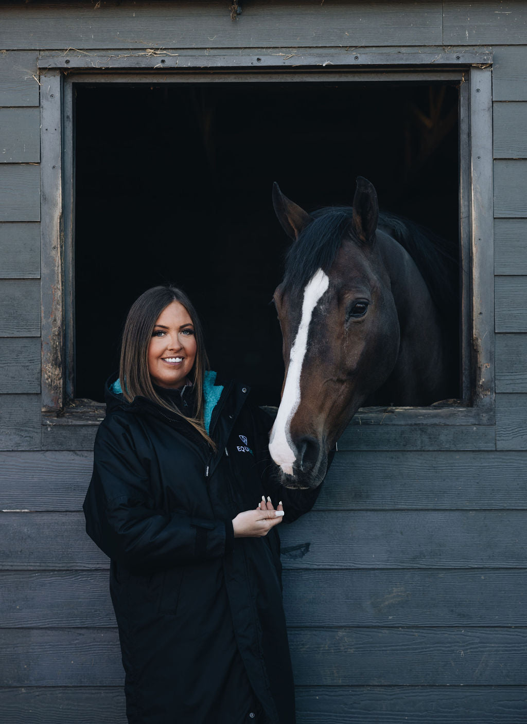 EQUIDRY Waterproof Riding Coat: Woman in Black/Turquoise Equestrian Jacket with Horse in Stable.