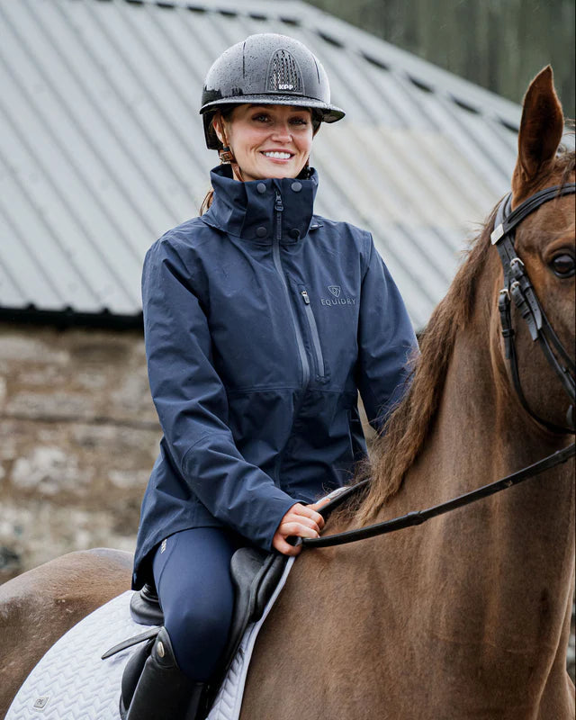 Equidry IRIS Waterproof Riding Jacket for Women in Navy. Woman riding a horse in a navy riding jacket and helmet.