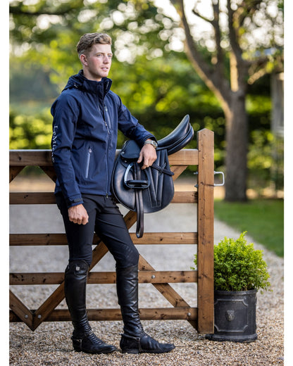 LeMieux Mens Elite Navy Soft Shell Equestrian Jacket. Man standing next to fence, saddle, and boots.