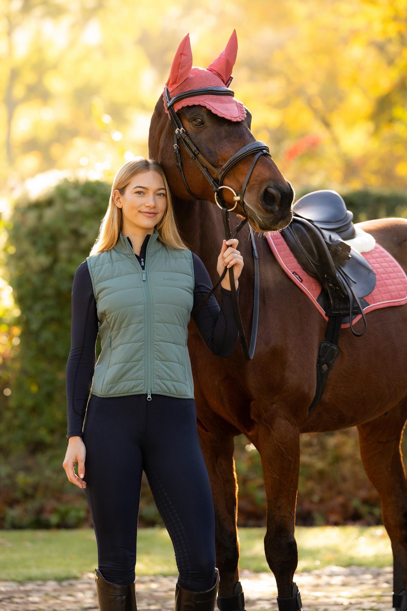 LeMieux Lily Lightweight Gilet Aloe: Woman in green gilet posing with brown horse and pink saddle pad.