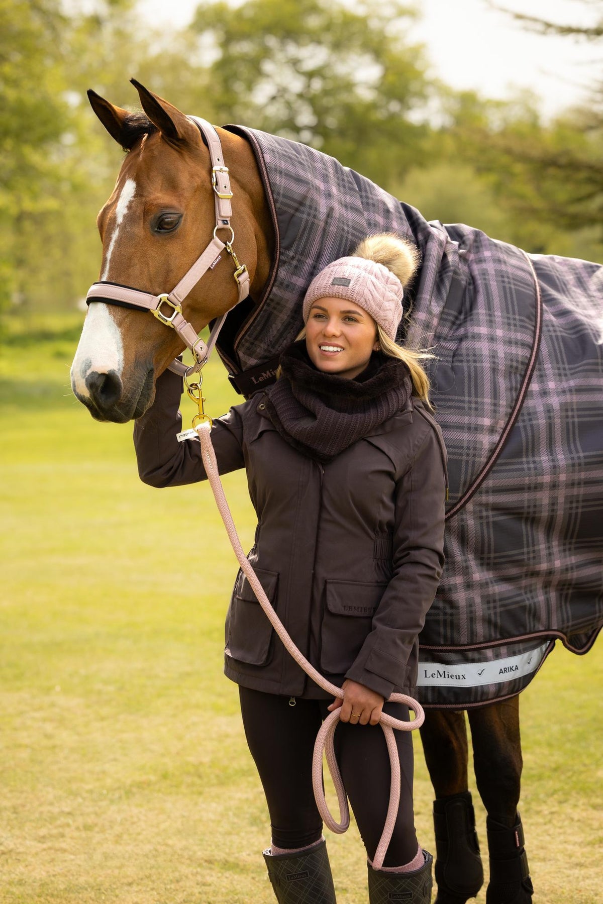Equestrian woman in LeMieux cocoa waterproof coat, pink hat, with horse & plaid blanket.