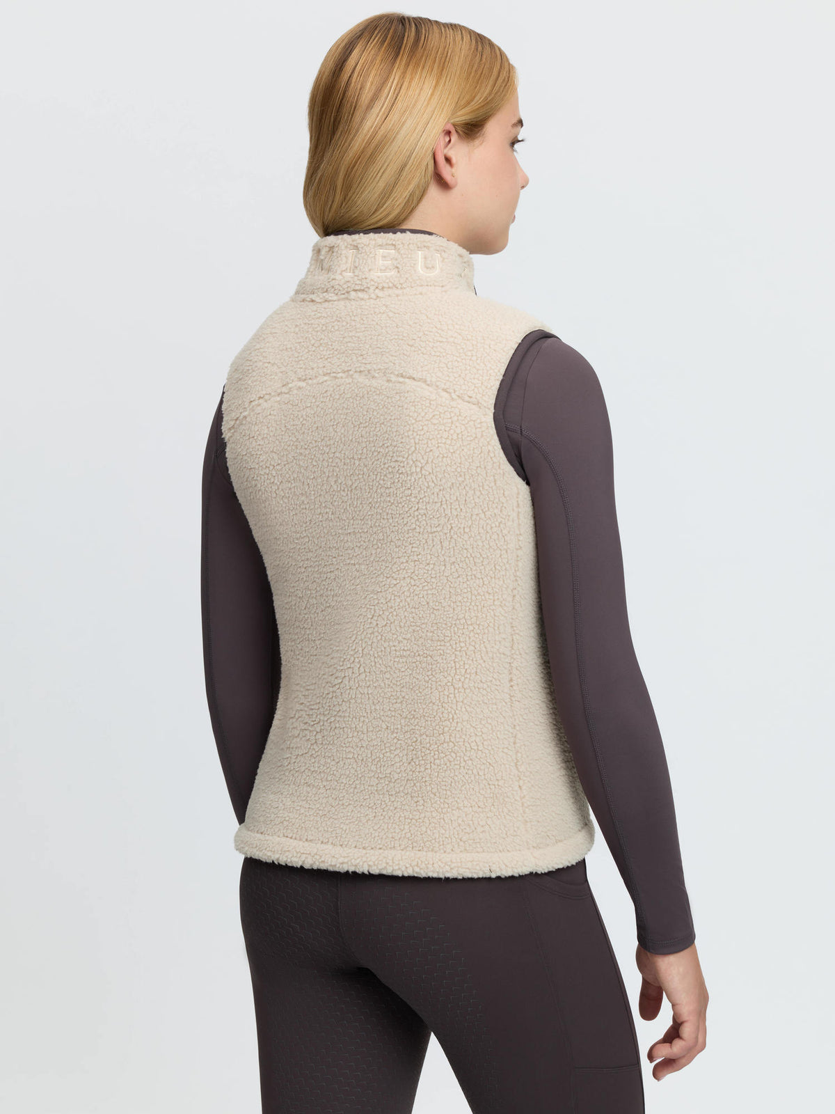 Young rider wearing a beige LeMieux teddy fleece gilet with a dark long-sleeved top and patterned leggings.