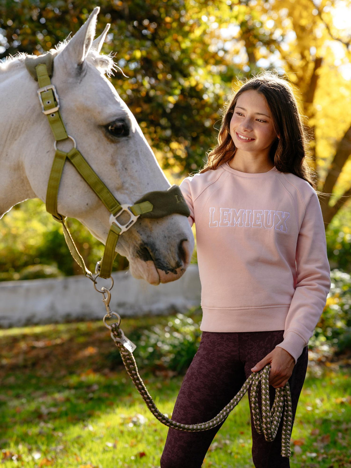 Young rider in a pink LeMieux varsity crew sweatshirt with a white horse outdoors