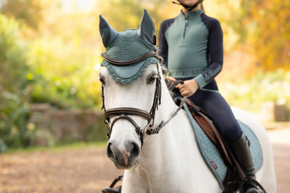 White horse wearing teal LeMieux fly hood and matching saddle pad, rider in equestrian attire