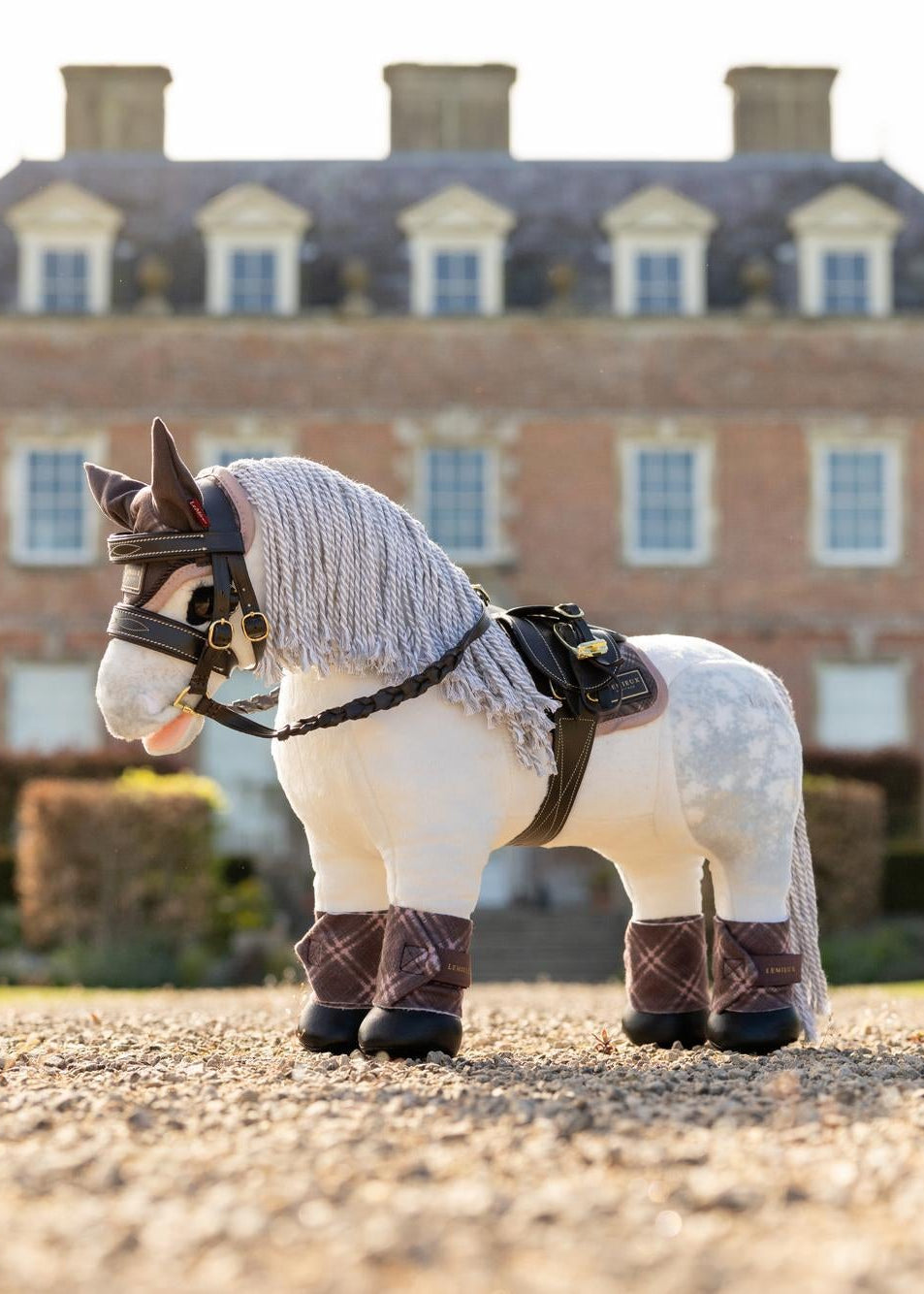 LeMieux Toy Pony Heritage Pad Cocoa - A plush white toy horse with grey spots, wearing a saddle pad and bridle, in front of a grand house.