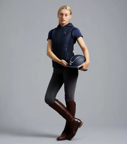 Equestrian woman wearing PREMIER EQUINE navy riding gilet and brown boots holding helmet.
