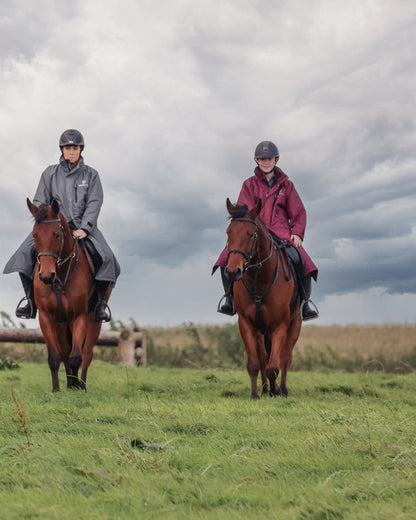 Two riders on horses wearing EQUIDRY waterproof riding coats (plum and grey). Horse riding in a field under cloudy sky.