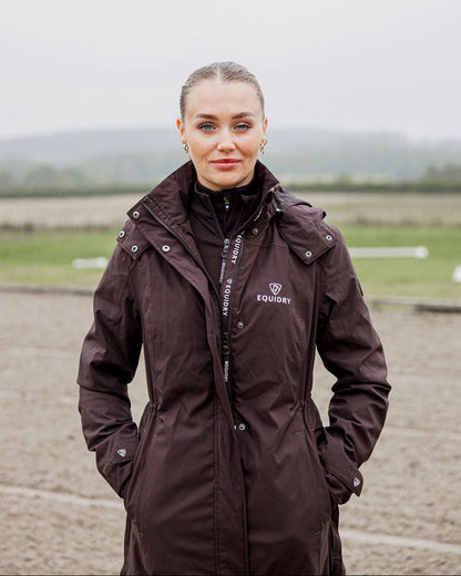 woman wearing brown waterproof tailored riding coat with Equidry branding. Long coat dark brown
