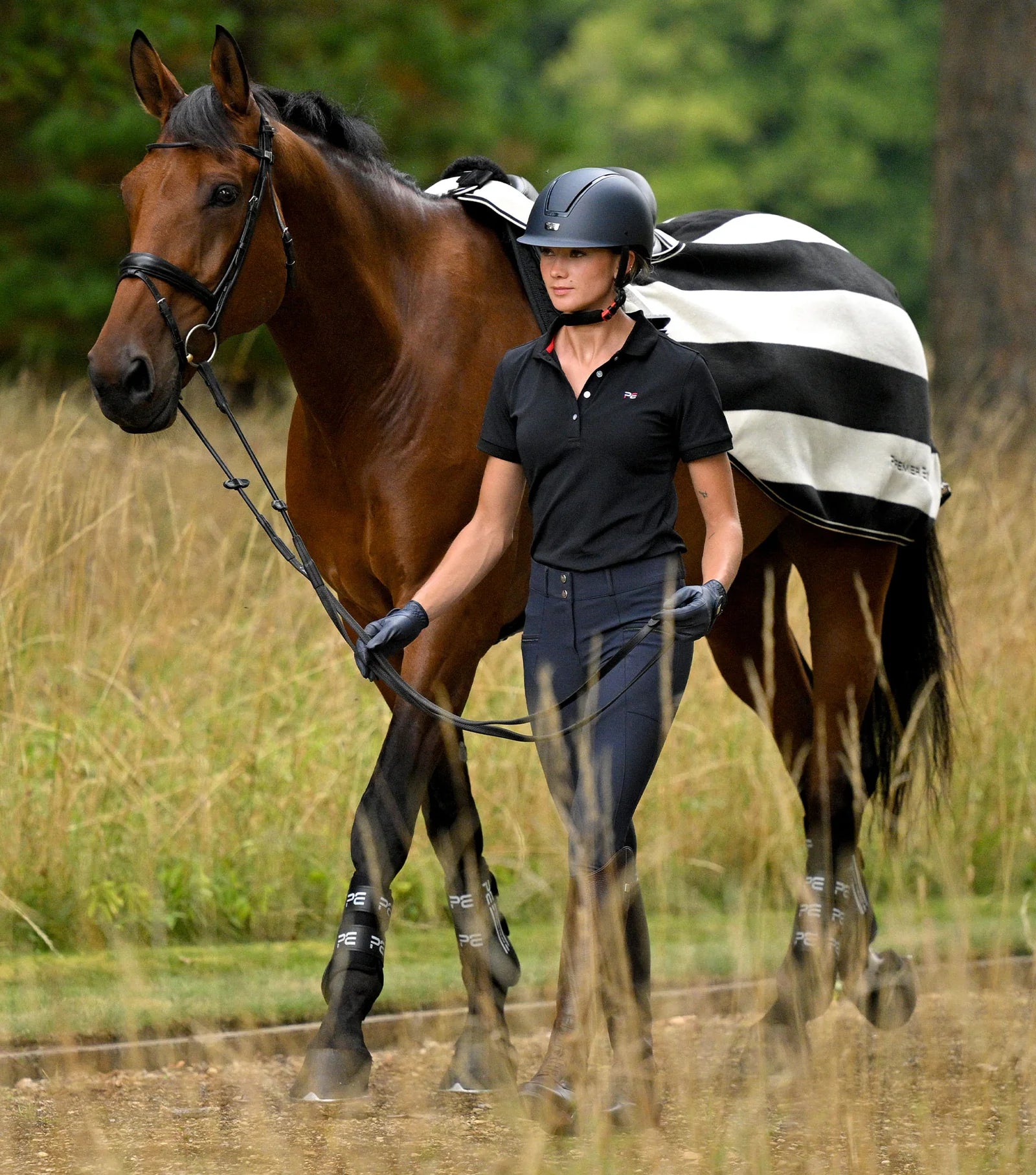 Equestrian: Woman leading horse with Premier Equine Vellus Fleece Exercise Rug in Black/White. Horse wear, riding.