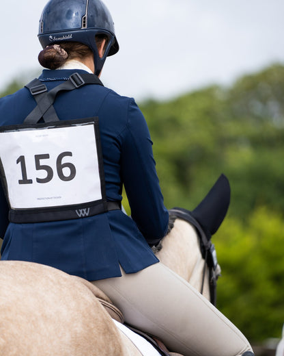 Horseback rider wearing a WOOF WEAR event number bib with number 156. Equestrian competition attire.