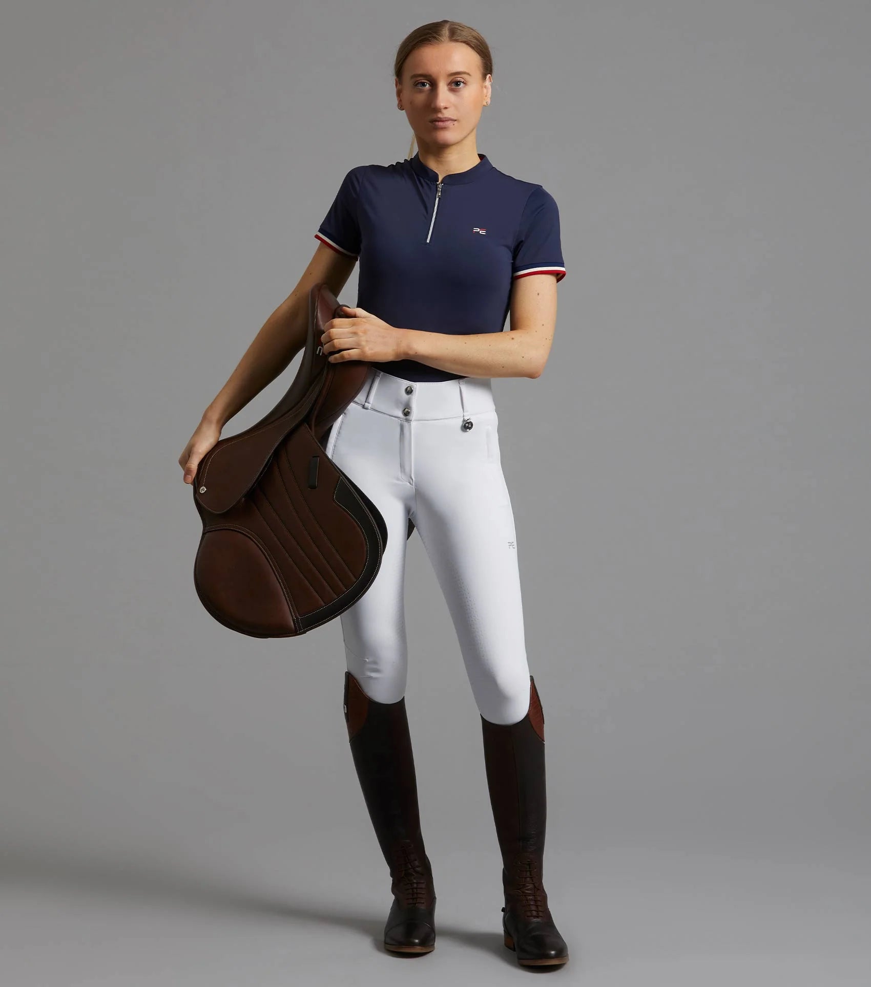 Equestrian: Woman in Premier Equine Navy Riding Top, White Breeches & Boots Holding Saddle