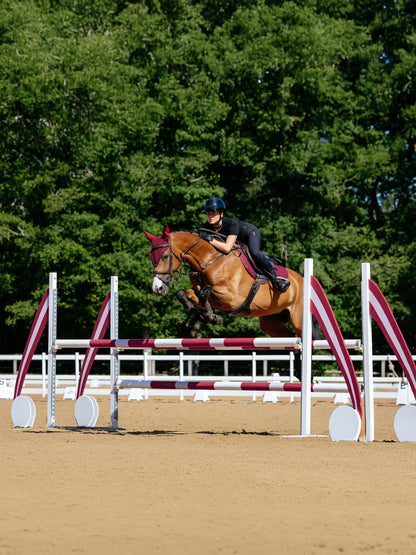 Horse jumping obstacle with LeMieux Cotton Close Contact Square burgundy pad from Equiflair, rider in black. Equestrian sports.