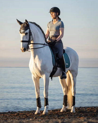 Equestrian in LeMieux Earth Dressage Square Ocean Saddle Pad on white horse by the sea. Sustainable horse riding gear from Equiflair.