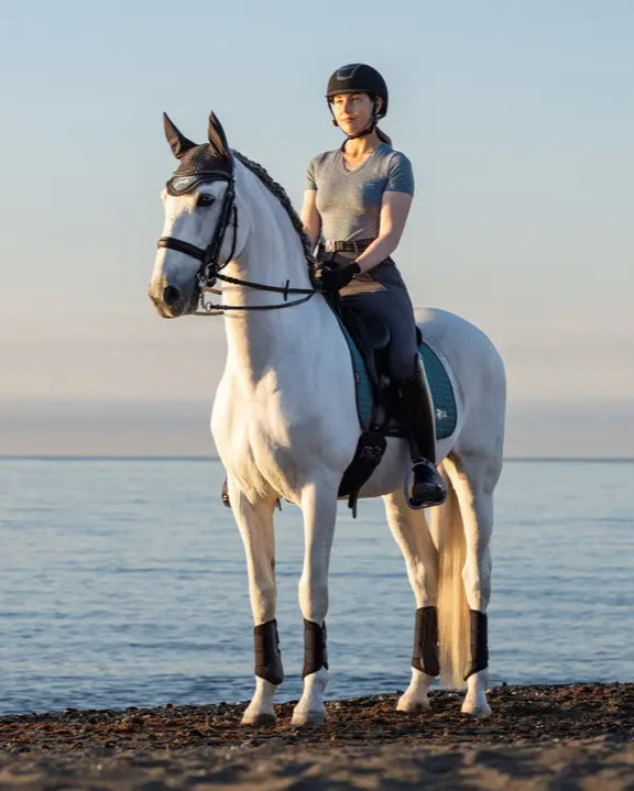 Woman rider on white horse with Lemieux Earth Dressage saddle pad. Equestrian dressage on a beach. Horse riding equipment.