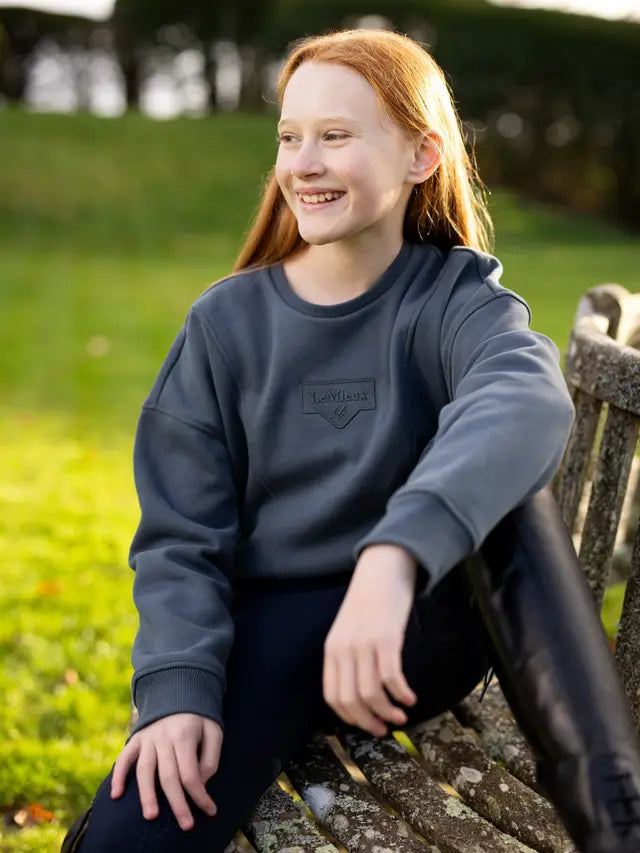 LeMieux Young Rider Clea Crew Sweatshirt in Petrol Blue. Girl wearing stylish equestrian sweatshirt on bench, Equiflair store.