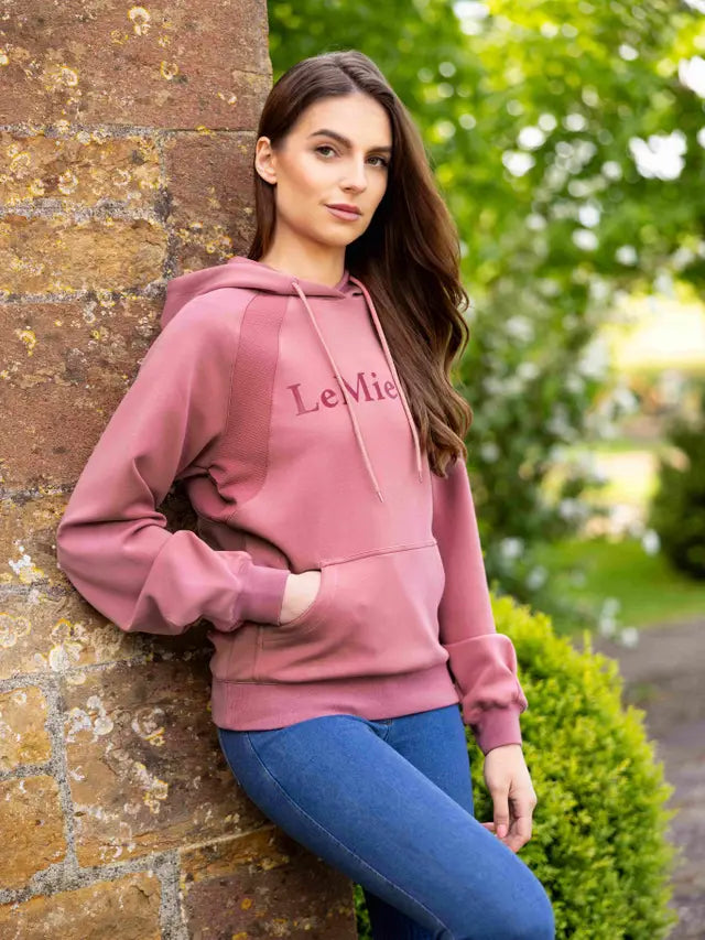 LEMIEUX NADINE HOODIE PEONY: Woman in comfortable pink hoodie & jeans, leaning against a brick wall outdoors.