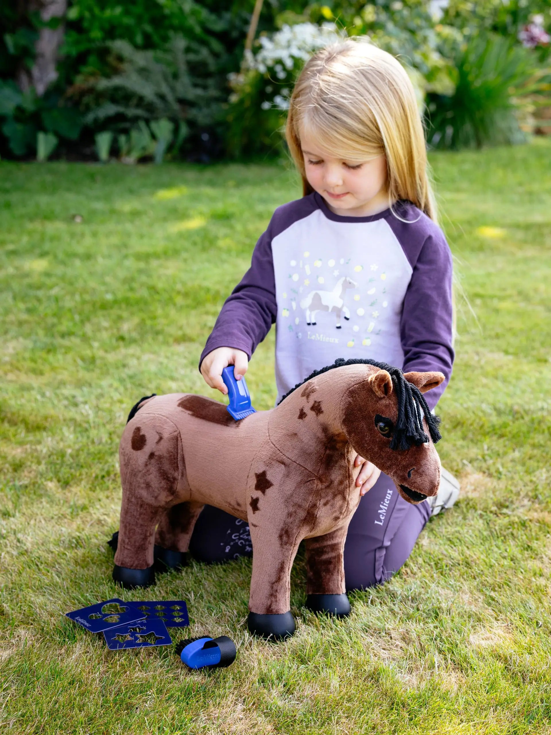 Child grooming LEMIEUX Toy Pony Gracie with brush. Horse role play with LeMieux toy horse and grooming kit on green lawn.