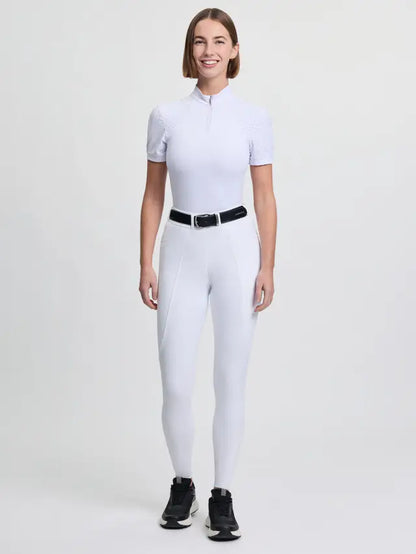 Woman wearing white LeMieux Brilliance Knee Grip Breeches and short-sleeved riding top, black belt, and black sneakers.