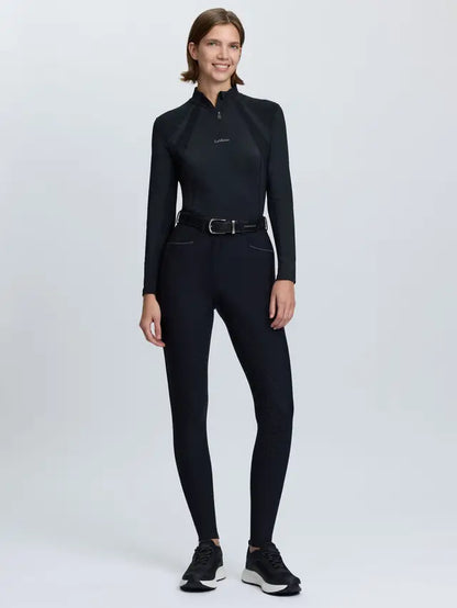 Woman wearing LeMieux black tall fit equestrian breeches and long-sleeve top, standing against a white background.