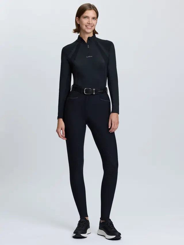 Woman wearing LeMieux black tall fit equestrian breeches and long-sleeve top, standing against a white background.