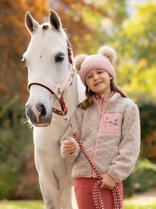 Young girl with white horse wearing LEMIEUX MINI THORA HEART FLEECE STONE jacket and pink beanie.