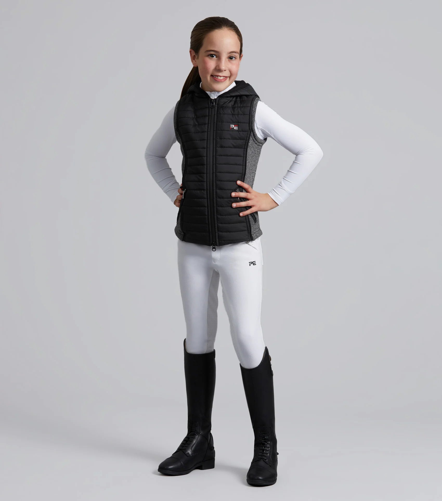 Girl in Premier Equine Black Hybrid Riding Gilet. Equestrian style with riding boots and white breeches.