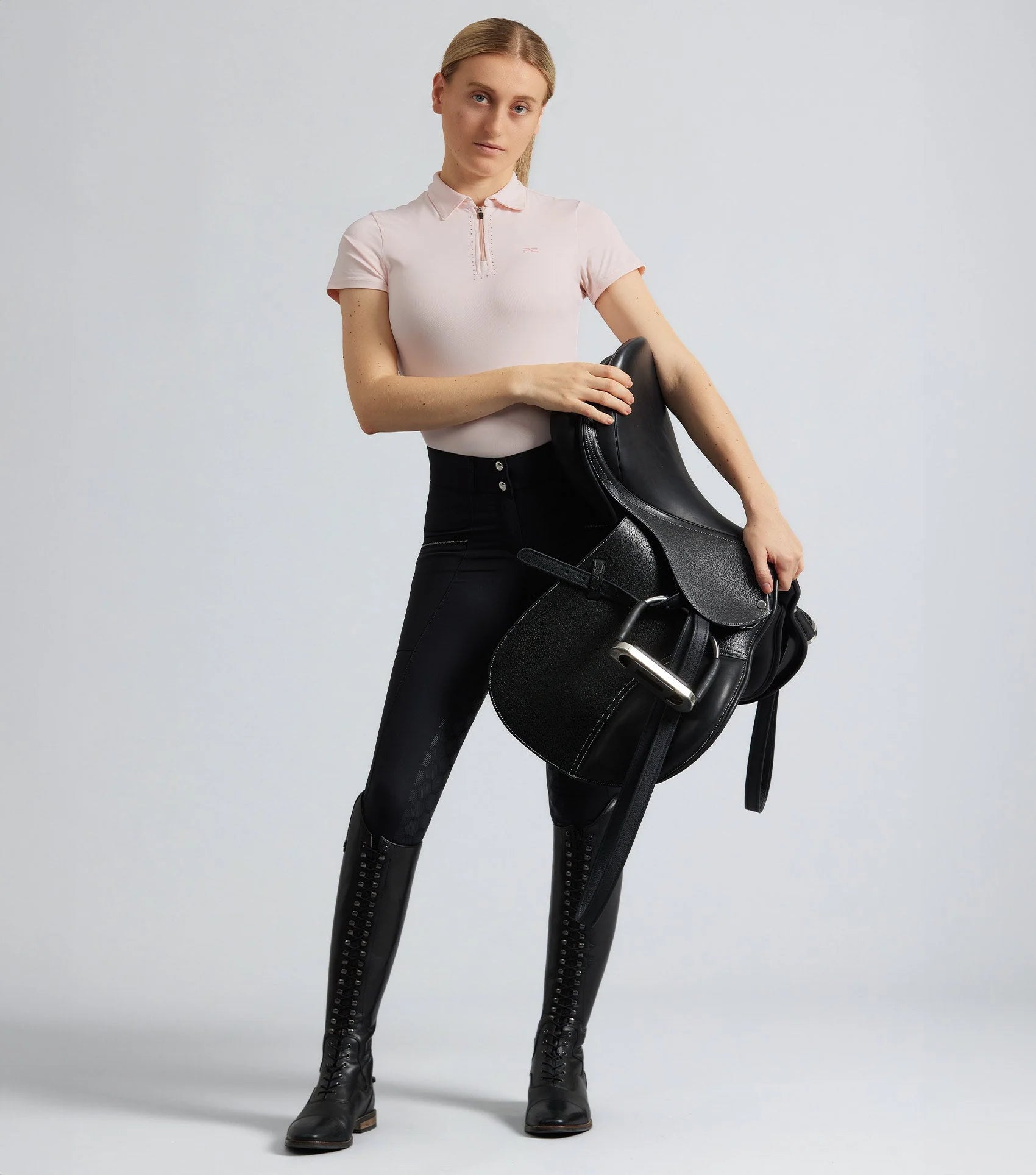 Woman wearing black Premier Equine riding breeches with full seat holding a horse saddle. Equestrian fashion.