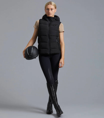 Premier Equine Ladies Quilted Gilet Black Equestrian Clothing. Woman in riding gear, holding helmet. Stylish equestrian apparel.