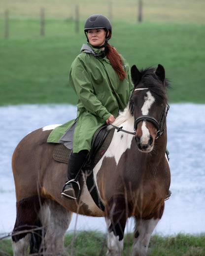 Woman riding a horse wearing a green waterproof Reincoat Lite poncho. Equestrian apparel.