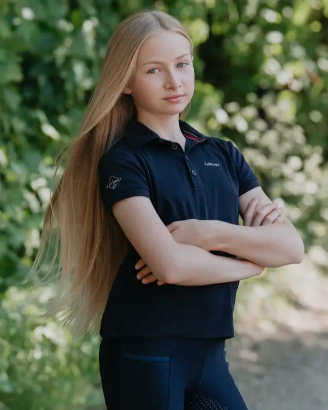 Girl in LEMIEUX Junior Pro Polo Shirt Navy, Equestrian Riding Apparel from Equiflair. Long blond hair, arms crossed.